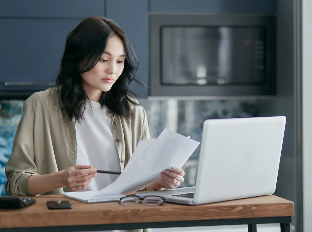 A focused woman sits at a kitchen counter reviewing documents beside an open laptop in a home office setting.