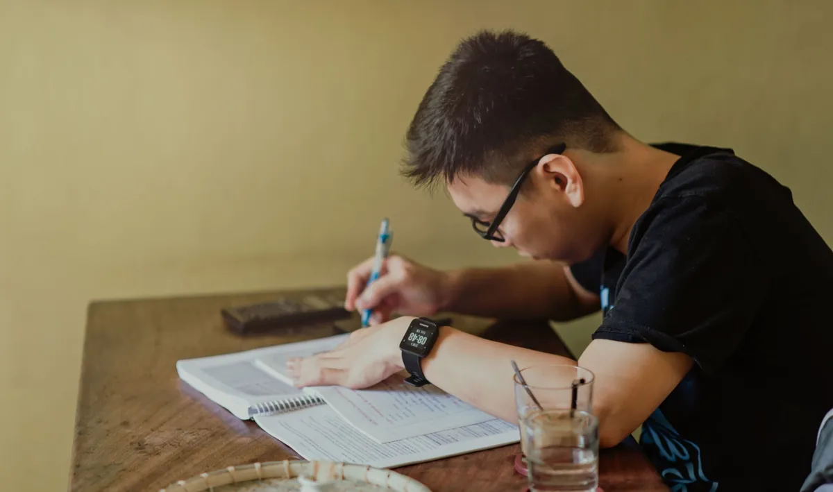 A young man sits at a desk indoors, concentrating as he writes notes while studying.
